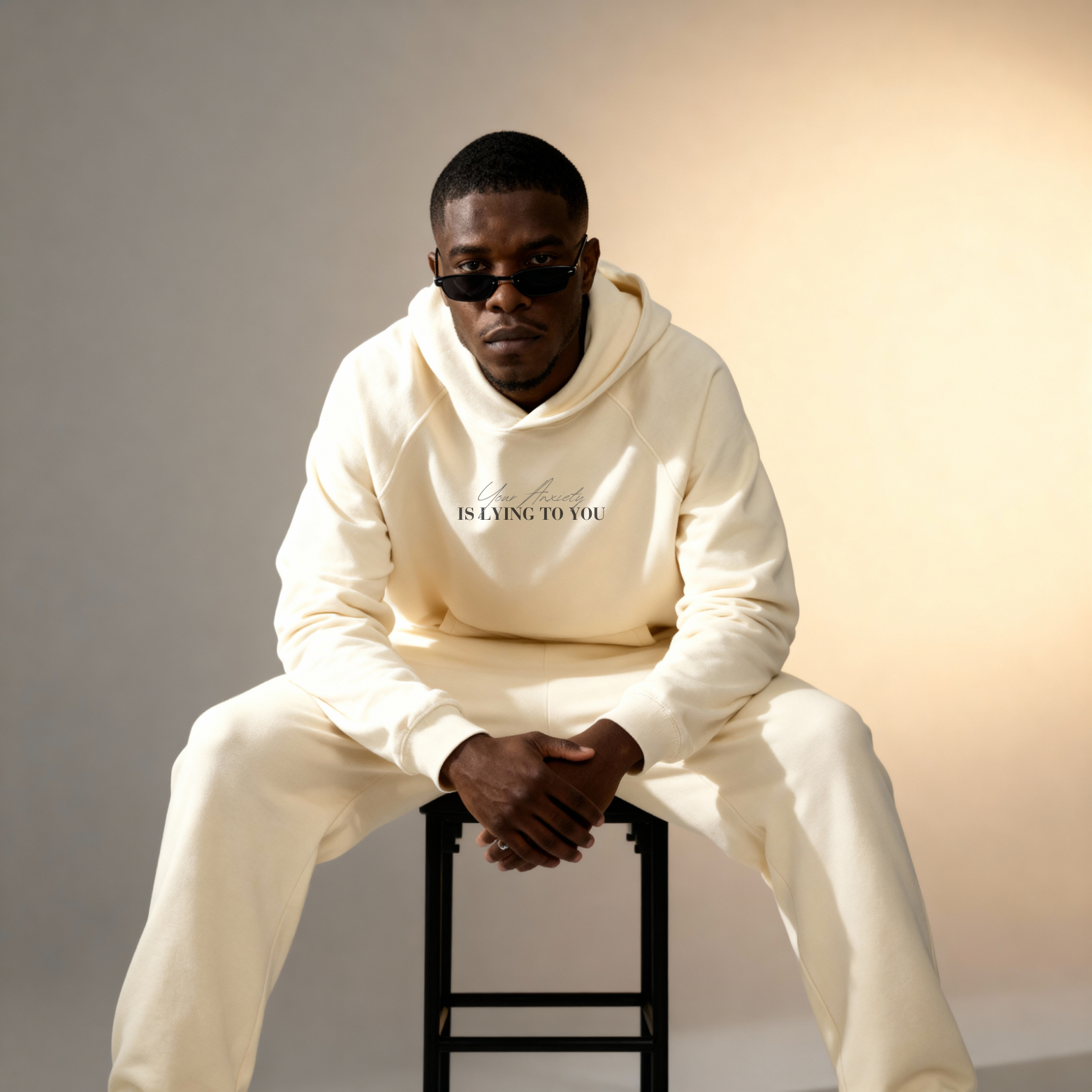 Man wearing a cream hoodie with text 'Your Anxiety IS LYING TO YOU' and pants sitting on a stool against a gradient background