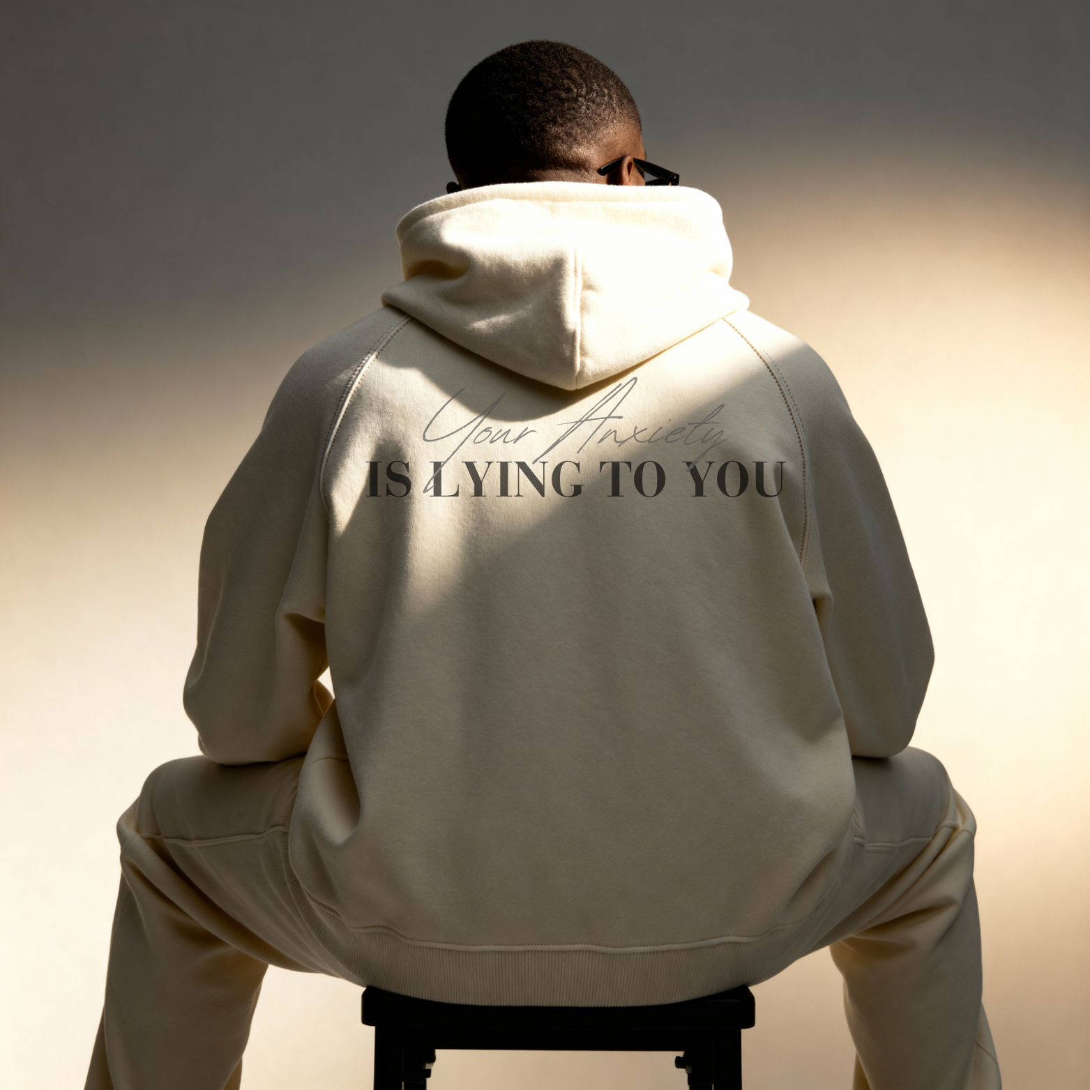 Person wearing a beige hoodie with text on the back of the Hoodie 'Your Anxiety IS LYING TO YOU' on a white background, sitting on a stool against a gradient background.